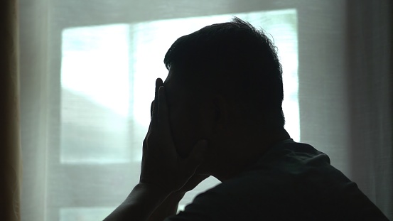 Silhouette of asian man sitting alone by window dark room , head in their hands,  feelings of sadness,depression,hopelessness ,Mental Distress Consept.