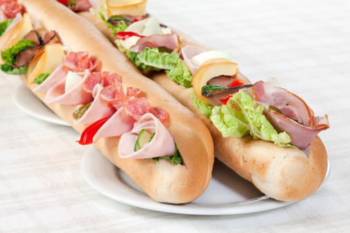 Two long sandwiches with ham, salami, eggs, cheese, pepper, lettuce and cucumber on three plates. Selective focus, shallow DOF.                                                                                                                                                                                                                                                                                                       