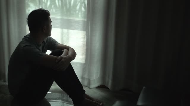 Asian man sitting alone by window dark room , head in their hands,  feelings of sadness,depression,hopelessness.