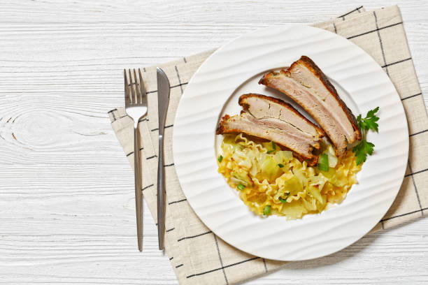 Haluski, fried cabbage, egg noodles, and butter served with roasted ribs on bone on white plate on white wooden table with cutlery, horizontal view from above, flat lay, free space