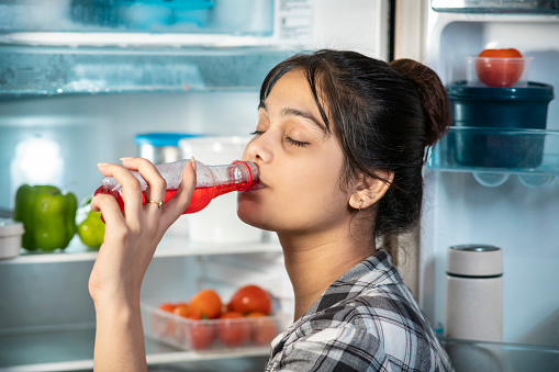 Close-up of young woman standing in front of open refrigerator and drinking soft drink from bottle at home in the summers.