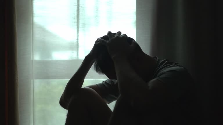 Silhouette of asian man sitting alone by window dark room , head in their hands,  feelings of sadness,depression,hopelessness.