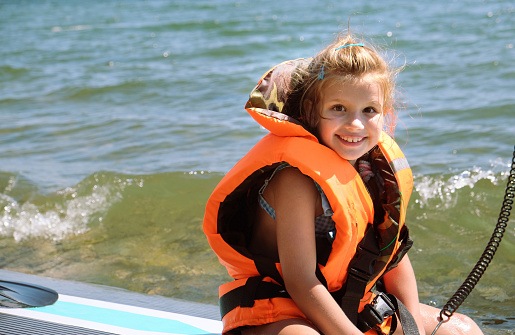 Portrait of happy smiling child girl with safety vest on the surf board, swimming board. Summer vacation leisure activity. Orange life jacket