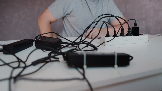 A man plugs many black electrical plugs into a surge protector. Many electrical wires are mixed up on a white table. Concept of safety violation, electric shock. Close-up