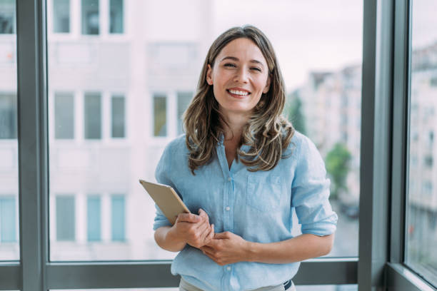 Confident businesswoman in modern office. Shot of successful young businesswoman standing in the office and holding digital tablet. Portrait of a beautiful smiling businesswoman standing in her office and looking at camera. professional woman smiling stock pictures, royalty-free photos & images