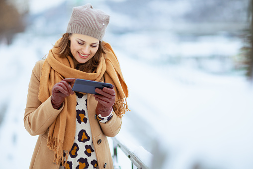 smiling stylish middle aged woman in brown hat and scarf in camel coat with gloves using smartphone app outside in the city in winter.