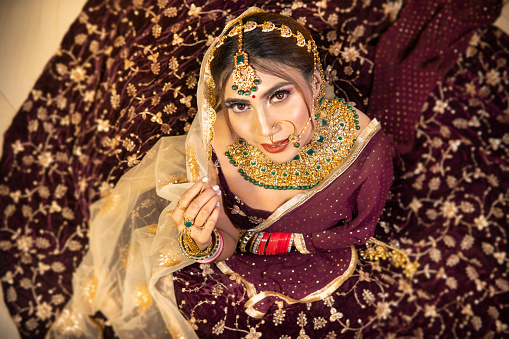 top viewp portrait of beautiful traditional Indian bride wearing lehenga and jewelry on her wedding day and looking at the camera with a smile.