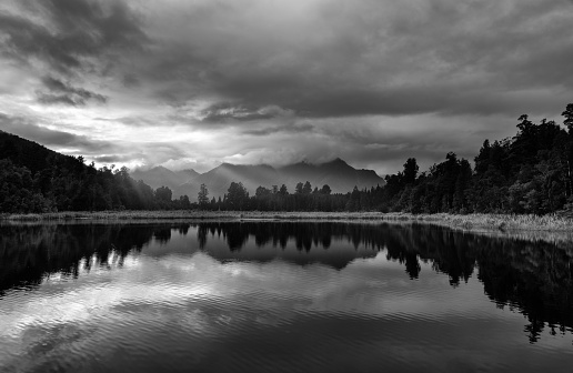 Dawn breaks over spectacular Lake Matheson in New Zealand. Heavy cloud drifts across the snowcapped peaks of the Southern Alps.