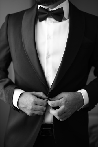 A young adult man wearing a formal tuxedo adjusting his black bow tie in a mirror, getting ready for a special event