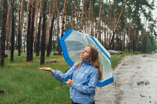 Young woman holding transparent blue umbrella outdoors in forest. Rainy weather day using umbrella. Woman with hand checking how long it will be raining. Forecast