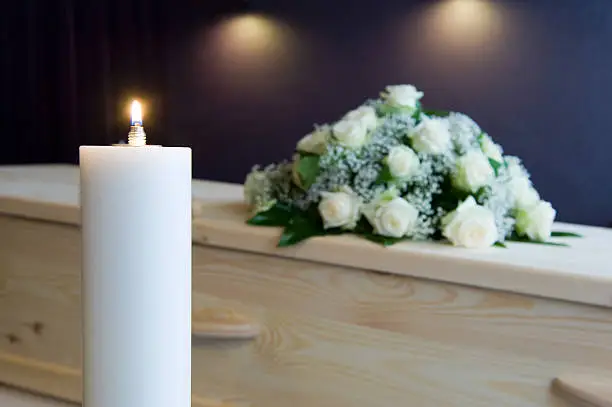 A burning candle with a coffin and a flower arrangement on the background in a mortuary