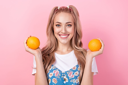 Portrait of toothy beaming pretty girl with ponytails wear t-shirt under dress arms hold two oranges isolated on pink color background.