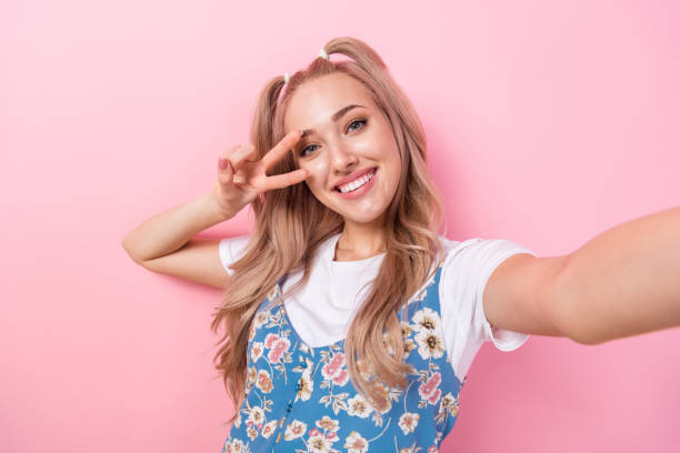 Portrait of cheerful optimistic girl with ponytails wear white t-shirt making selfie show v-sign isolated on pink color background stock photo