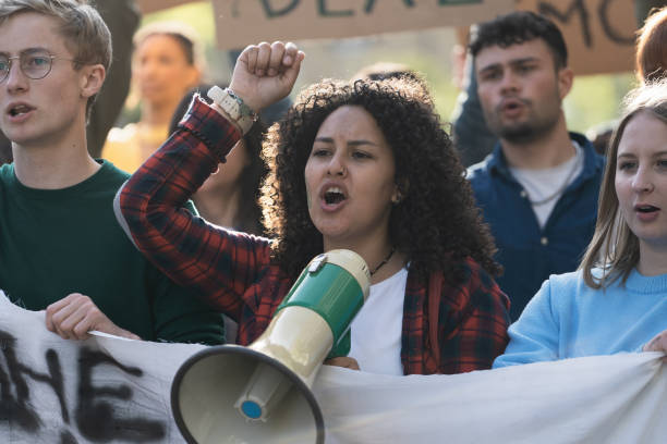 Student Activist Leading Protest with Megaphone Close-up of a young woman with curly hair, leading a student protest using a megaphone. The group, holding an unidentifiable banner, passionately chants slogans. protestor stock pictures, royalty-free photos & images