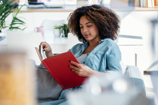 istock Pretty young woman reading a book while sitting on sofa at home. 1705630291