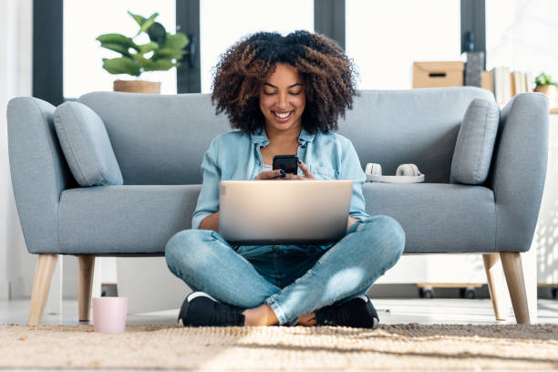 istock Pretty young woman using her mobile phone while working with laptop sitting on the floor at home. 1705630839