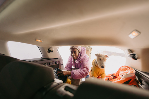 Photo of a young African American woman and her dog, who enjoy hiking and spending time in nature, unpacking backpacks from car trunk, as they have parked at the foot of the snowy mountain