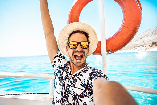 Happy tourist man enjoying summer vacation sailing on a boat in the sea - Cheerful laughing guy taking selfie portrait with smart mobile phone outside - Tourism life style and technology concept