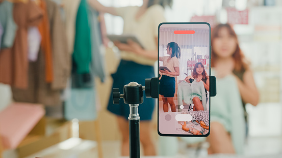Closeup of young Asian women live streaming on phone show new collection fashion clothes to customer at shop. SME small business e-commerce concept.