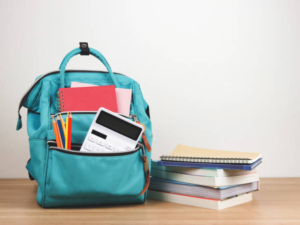 istock Front view of green backpack with school supplie, stack of books on wooden table and white  background with copy space. 1705762907