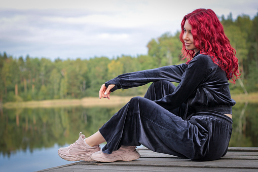 Portrait of a stylish young woman with red hair in a tracksuit on the river bank. Calmness and tranquility.