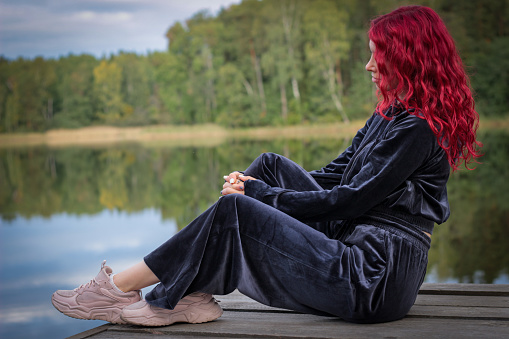 Portrait of a stylish young woman with red hair in a tracksuit on the river bank. Calmness and tranquility.
