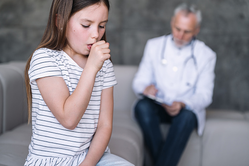 Selective focus on preteen girl patient feel bad and coughing while sitting on couch and blurred doctor on background. Medical appointment at home. Kid with influenza symptoms on pediatrician visiting