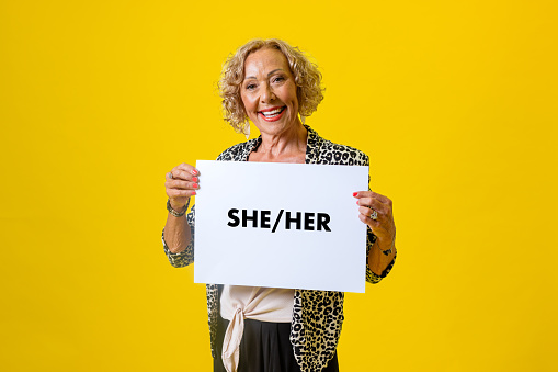Waist-up studio portrait of a senior female model looking into the camera with a cheerful expression, showing her teeth. She is holding a white sign displaying the words she and her on it.