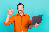 Portrait of satisfied cheerful man dressed orange shirt hand hold laptop showing okey approve isolated on turquoise color background