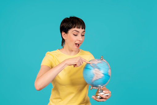 cheerful woman in yellow t-shirt pointing finger at earth globe choosing vacation spot isolated on blue background Passenger traveling abroad. what you choose. journey, education concept