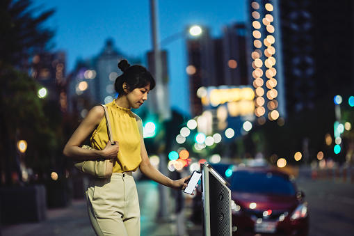 In the nighttime, an Asian woman in the city is using her mobile phone to make a payment for parking, embracing the concepts of digital wallets and diverse payment methods.