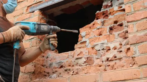a worker in a black T-shirt and blue jeans with a powerful drill in gloved hands hollows out a hole for a window in a red brick wall from the street side, working with a jackhammer on brickwork