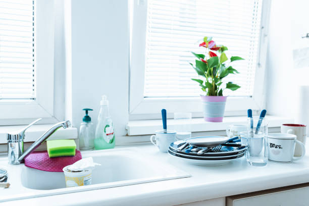 stack dirty plates with forks, cups, stand near kitchen sink, in kitchen with white facades. Unwashed dishes in pile on kitchen table near sink