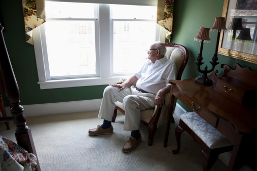 Contemplative senior man (80s) sitting in chair at home, looking out the bedroom window.