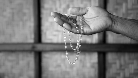 Person holding a rosary beads in hand in black and white background with light. Praying rosary concept, catholic symbol of devotion to Mother Mary.