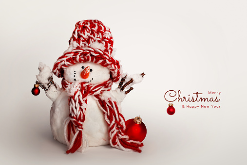A snowman made of cotton wool in a red hat and scarf holds a Christmas red ball. Postcard for congratulations on the New Year and Christmas holiday.