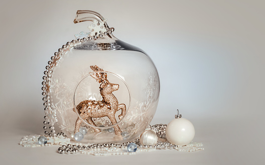 A Christmas toy deer stands in a glass apple on a blue background. There are snowballs, streamers, beads, balls, tinsel lying around.