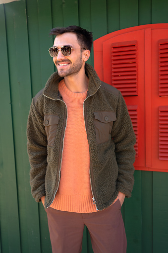 Stylish man stands next to wooden wall. Fashionable male posing near wooden house with green walls and red window shutters. Handsome male in dark sunglasses has confident smile