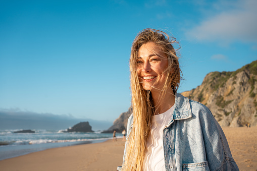Woman with happy smile on beach. Portrait of female smiling at sea shore with huge rocks on background. Blond woman stand on sandy beach and has radiant smile on her face. Vacation Traveling concept