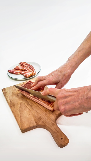Senior woman hands, cutting pork bacon slices with Santoku knife, on wooden chopping board, high resolution stock photo.