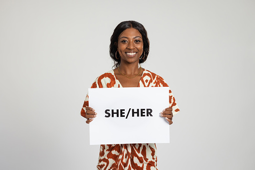 Waist-up studio portrait of a teenage model looking into the camera with a cheerful expression. She is holding a white sign displaying the words she and her on it.