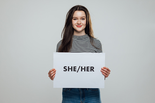 Waist-up studio portrait of a teenage model looking into the camera with a cheerful expression. She is holding a white sign displaying the words she and her on it.