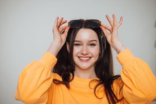 Studio portrait of a teenage female model looking into the camera while smiling. She is wearing a bright yellow jumper and is holding a pair of sunglasses on her head.
