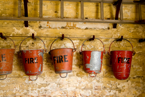 A row of old damaged fire buckets in a dark room. The handles on the bottoms are to make it easier to hurl the contents over flames.