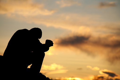 A silhouette of a man praying at sunset. Model is handsome Caucasian male in his 30s with unrecognizable side view. Model is folding his hands and bowing his head. Themes include meditation, spirituality, balance, freedom, vitality, hope, hurt, problems, addiction, struggle, anguish, help, god, healing, recovery, perseverance, coping, courage, strength, men, and asking for help. 