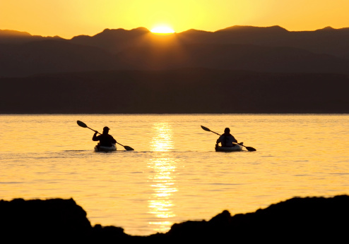 Two sporty outdoors people kayaking or canoeing on the sea, silhouetted as they paddle into the sun setting behind the mountains. See copy space version...