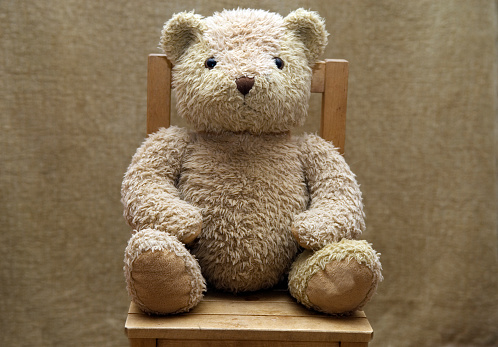 Old scruffy toy teddy sitting on child's wooden chair.