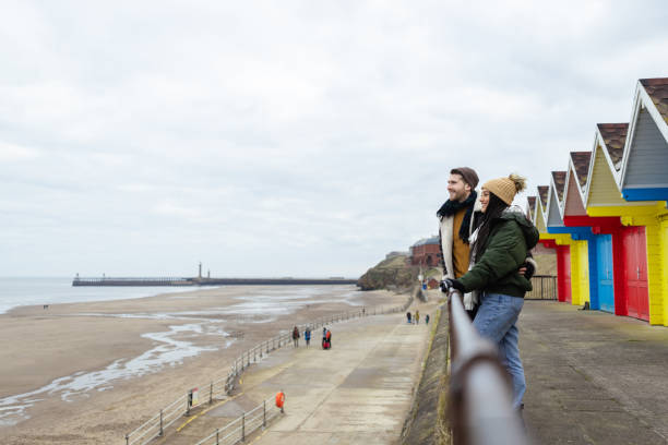 Side view of a mid adult couple enjoying walking along the pathway by the coast together. It's cold outside so they are wrapped up in warm clothing in Whitby, North Yorkshire. They have stopped to look at the view.