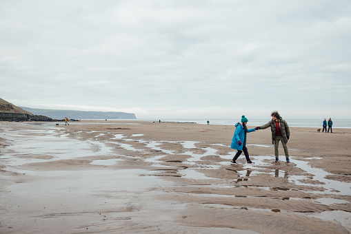 Wide view of a mid adult couple enjoying walking along the beach at the coast together. It's cold outside so they are wrapped up in warm clothing in Whitby, North Yorkshire.