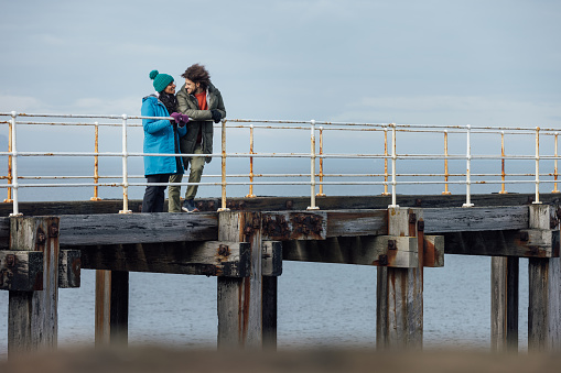 Wide view of a mid adult couple enjoying walking along the pier at the coast together. It's cold outside so they are wrapped up in warm clothing in Whitby, North Yorkshire. They have stopped to look at the view from the pier.